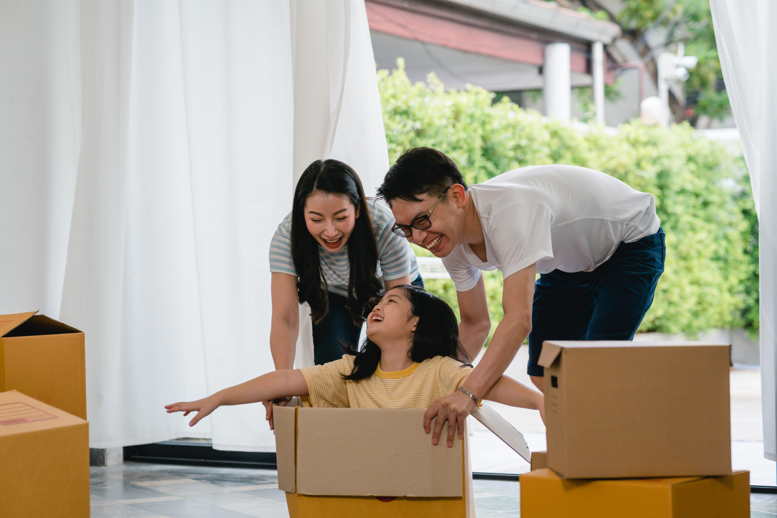 Happy Asian young family having fun laughing moving into new home. Japanese parents mother and father smiling helping excited little girl riding sitting in cardboard box. New property and relocation.